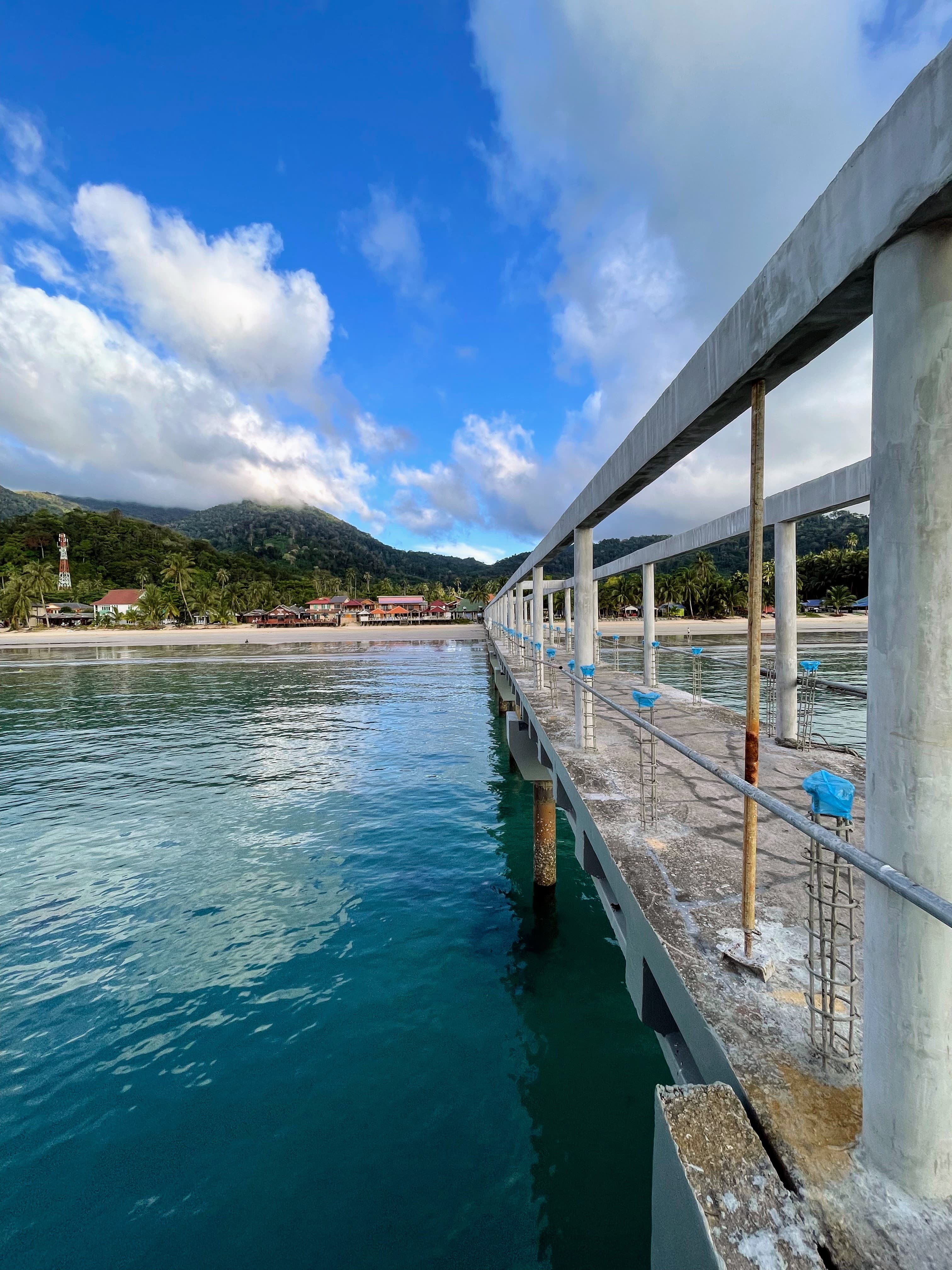 A concrete jetty under construction extending over shallow water toward a village and forested hills on Tioman Island
